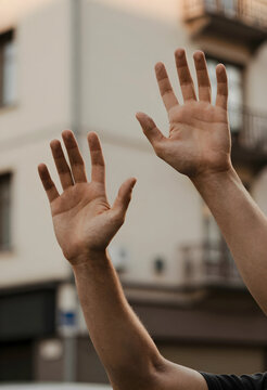 Raised hands with open palms in a gesture of greeting or surrender against a blurred urban background, the arms extend upwards against a softly blurred background of an urban building