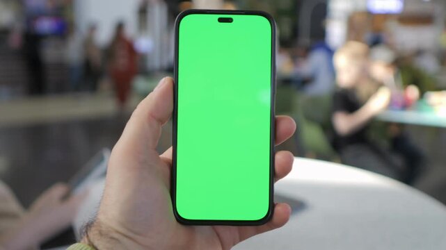 Hand holds phone with green screen. Background blurred cafe scene. Person focused on device. Ideal for app demos or digital content. Realistic indoor lighting
