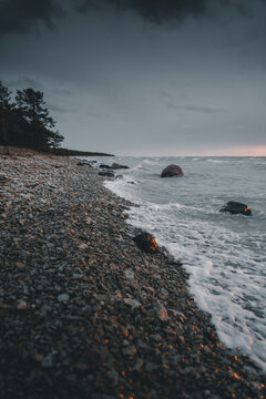 View of Saxby rannik featuring a rocky shoreline with waves crashing against stones under a dramatic dark cloudy sky at sunset in Saxby, Laane maakond, Estonia.