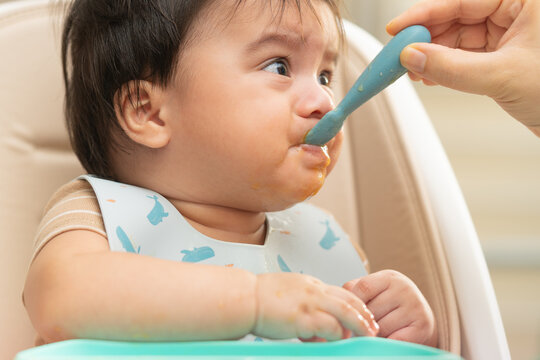 Baby being fed with a silicone spoon in a high chair during complementary feeding. Infant nutrition, parenting and childcare concept.