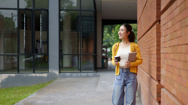 Young asian female student with backpack smiling while holding a book and a takeaway coffee cup, walking outside a modern building university campus