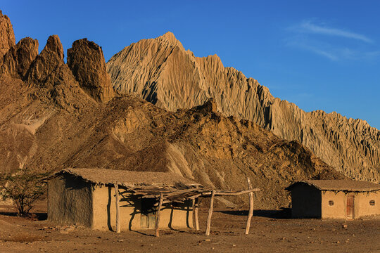View of traditional mud houses nestled against the rugged, sunlit rock formations of the Hingol landscape in Awaran, Balochistan, Pakistan.