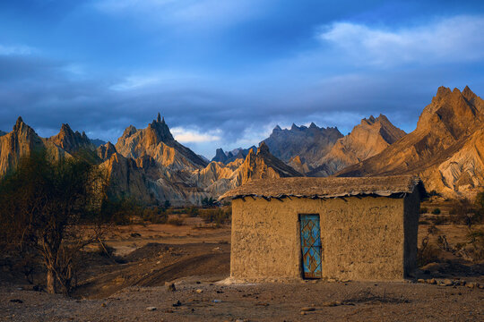 View of a small mud-brick house with a blue door set against the dramatic, jagged peaks of the Hingol mountains in a desert landscape in Awaran, Balochistan, Pakistan.