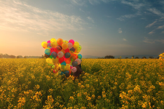 View of a person holding a large bunch of colorful balloons in a vibrant field of yellow mustard flowers during a golden sunset in Jhelum, Punjab, Pakistan.