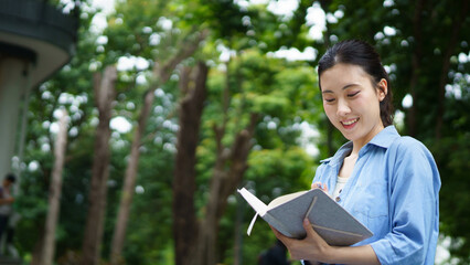 Fototapeta premium Education, university and woman in campus, Young asian woman smiles widely while tacking note. She standing in campus with her ready. Enjoying education outdoors with textbook