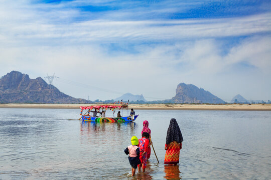 View of people wading in the shallow water of the Chenab River with a colorful boat in the background and mountains under a blue sky in Chiniot, Punjab, Pakistan.