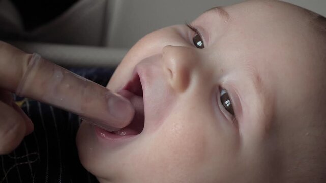 Mother or father gently massaging and cleaning gums and first milk teeth of newborn infant using special silicone finger toothbrush. Oral hygiene and to soothe teething pain