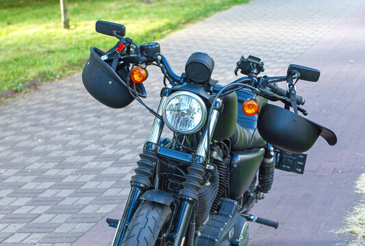 A front view of a black cruiser motorcycle parked on an outdoor paved driveway. Visible are helmets hanging from the handlebars, a round headlight, mirrors, and amber turn signals