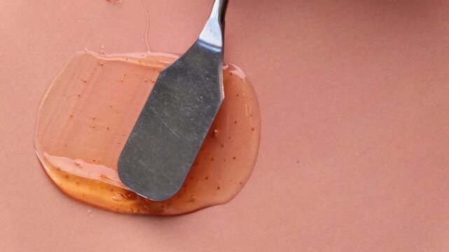 Exfoliating orange scrub gel texture smear by palette knife on matching background. A macro slow-motion shot captures a metallic palette knife smoothly spreading a vibrant orange exfoliating scrub gel