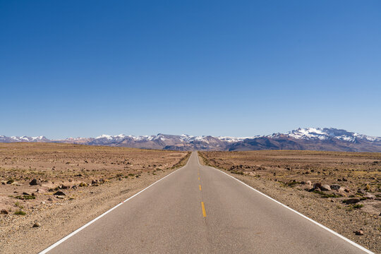View of the Mirador de los Andes road stretching through the arid high-altitude landscape towards snow-capped mountains under a clear blue sky in Caylloma, Arequipa, Peru.