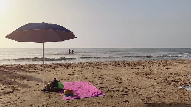 Beach umbrella stands in the sand with a backpack containing a towel and water bottle underneath, while a hat with sunglasses and flip flops rest beside it. Three women are seen in the sea