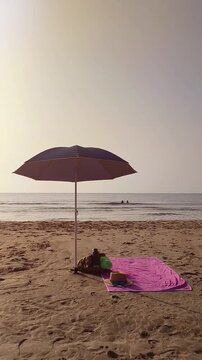 Vertical video of a beach umbrella planted in the sand with a backpack containing a towel and water bottle underneath, beside a hat and flip flops. Three women are visible in distance 