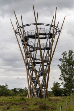 View of View Tower D'n Flaestoren, a modern wooden and metal observation tower with a spiral staircase, set in a grassy landscape under a cloudy sky in Lage Mierde, Noord-Brabant, Netherlands.