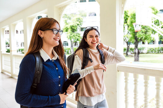 Two students with backpacks walk and laugh along a bright campus corridor with arched railings and greenery outside.