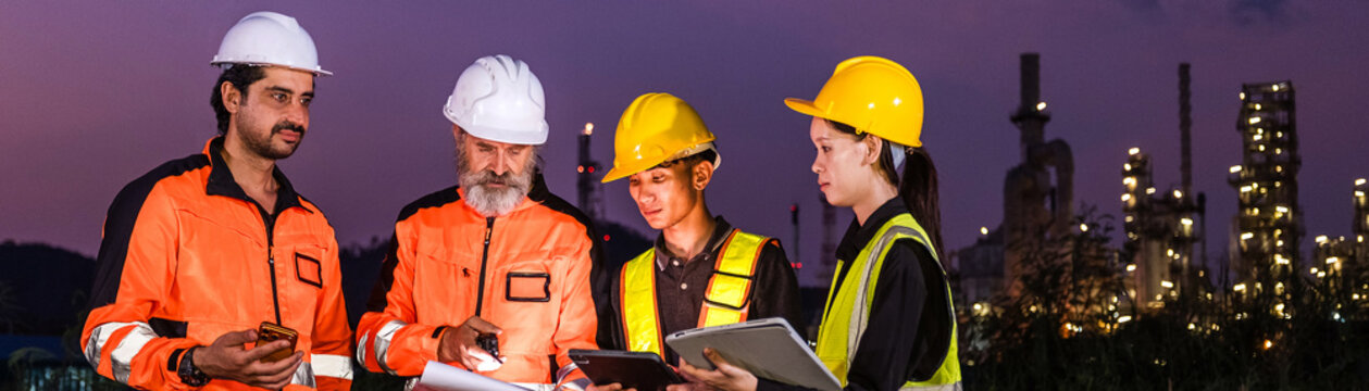 Industrial workers in safety gear review plans at a refinery site at dusk, wearing helmets and high-visibility clothing with plant lights glowing in the background.