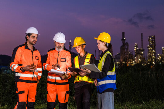 Industrial workers in safety gear review plans at a refinery site at dusk, wearing helmets and high-visibility clothing with plant lights glowing in the background.