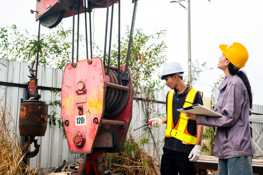 Construction workers inspect a red crane hoist and hook at a jobsite, using a clipboard and wearing safety gear beside metal fencing and overgrown weeds.
