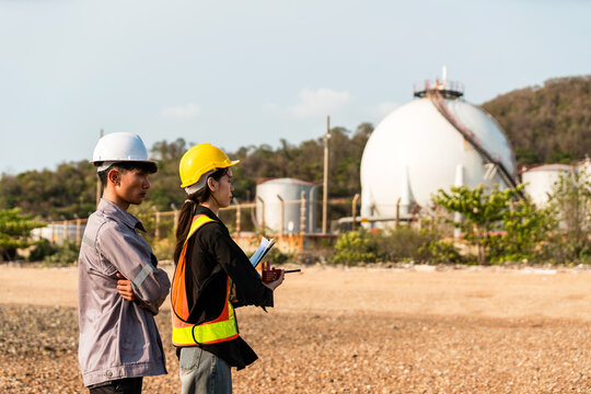 Two engineers in hard hats and safety vests inspect an industrial energy site with spherical gas storage tanks.