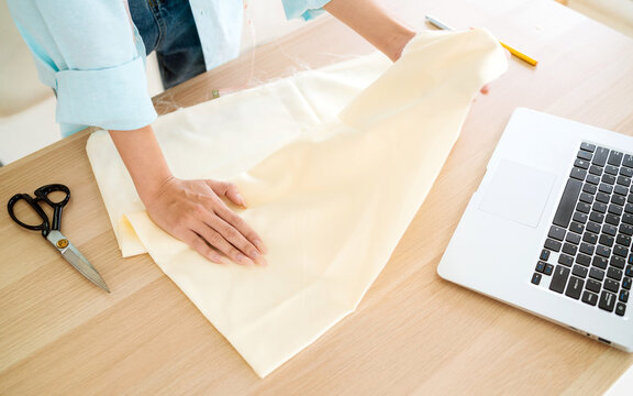 Person folding cream fabric on a worktable beside a laptop and scissors, preparing a sewing project in a home studio