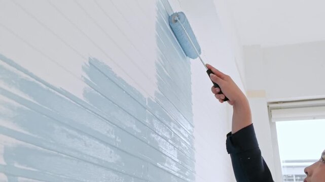 Hand using a paint roller to apply light blue color on an interior wall during a home renovation near a window.