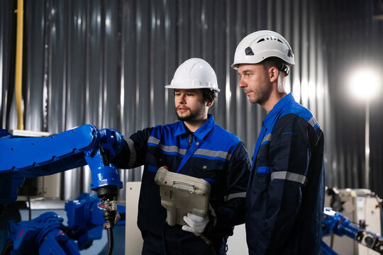 Two professional industrial engineers in safety hard hats programming an automated robot arm using a teaching pendant in a modern Industry 4.0 smart manufacturing factory facility.