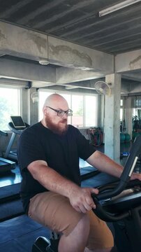 Bearded man with glasses pedals a stationary bike in a modern gym, focused on cardio amid treadmills and weights, with daylight streaming through windows.
