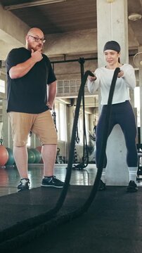 Woman training with battle ropes in a gym while a coach supervises; industrial interior, focused workout