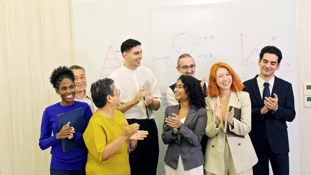 Diverse team in an office applauds during a presentation, standing before a whiteboard with charts and diagrams, celebrating results and teamwork