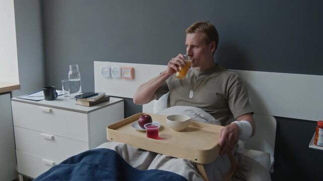 Young adult Caucasian man with bandaged arm lying in hospital bed, drinking orange juice from breakfast tray. Footage for healthcare, rehabilitation, or military recovery ads