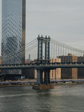 View of the Manhattan Bridge with its blue steel structure and suspension cables, set against a backdrop of modern glass skyscrapers and residential buildings in New York, New York, United States.