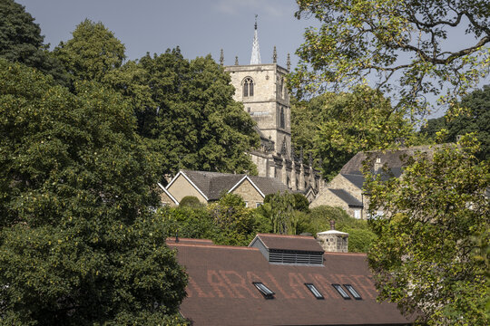 View of the historic St John the Baptist Church tower rising above lush green trees and stone rooftops in Knaresborough, England, United Kingdom.