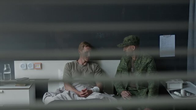Young adult Caucasian soldier lying in hospital bed with bandaged hand talking to middle aged Caucasian officer. Footage for veteran support and military healthcare marketing