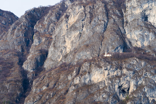 View of a small church perched on a rocky cliffside with rugged mountain terrain and sparse vegetation in Varenna, Lombardia, Italy.