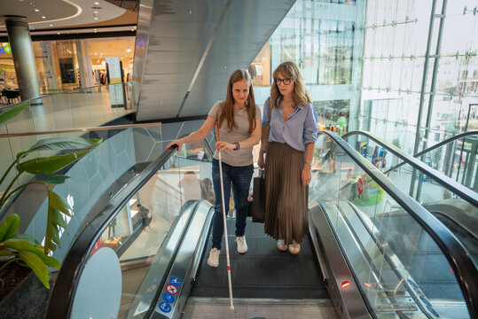 Visually impaired woman with female friend standing on an escalator in a modern shopping mall