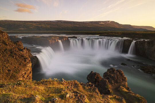 View of Godafoss Waterfall with cascading water flowing over rocky cliffs into a turquoise pool under a soft golden hour sky, Thingeyjarsveit, Iceland.