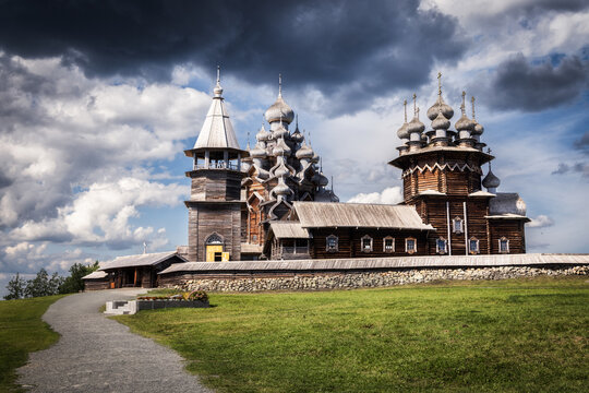 View of the historic wooden Kizhi Pogost with its iconic onion domes, bell tower, and surrounding stone wall under a dramatic cloudy sky, Petrozavodsk, Republic of Karelia, Russia.