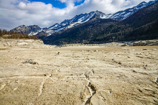 View of the cracked, parched bed of the dried-up Ceresole Reale lake during a drought, with snow-capped mountains in the background under a blue sky in Ceresole Reale, Piedmont, Italy.