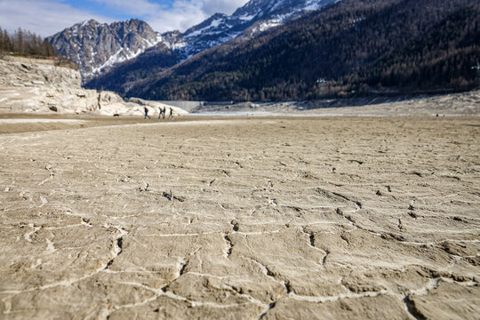 View of the cracked, dried-up bed of Ceresole Lake during a drought, with people walking in the distance near snow-capped mountains in Ceresole Reale, Piedmont, Italy.