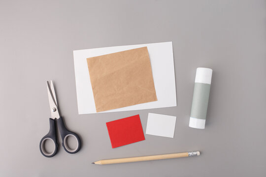 Scissors, glue stick, pencil, colored paper squares and blank white card on grey background. Flat lay craft supplies mockup with copy space. DIY paper craft and education concept.