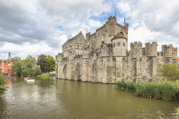 Obraz premium Gravensteen, a medieval castle in the city of Ghent in Belgium, Europe. Historic Stone Castle and Moat with Tourist Boat on a Cloudy Day, Reflecting Architectural Heritage