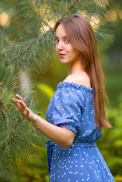 A young smiling woman strolls through a spring park in a good mood. A beautiful young girl with long light brown hair in a dress poses among plants, trees, and flowers.
