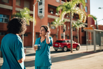 Fototapeta premium Two nurses enjoying a coffee break, talking outside a hospital building. They are wearing scrubs, representing medical staff