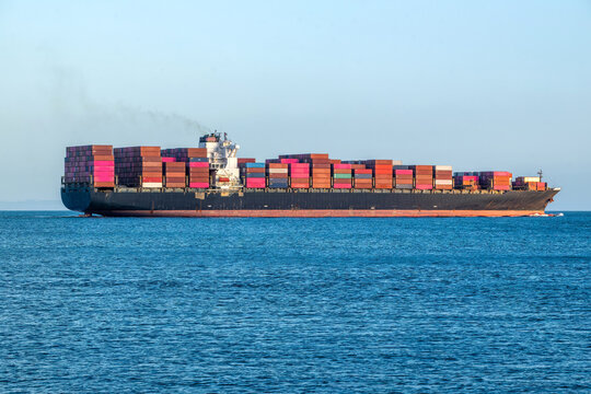 View of a large cargo vessel loaded with colorful shipping containers sailing on the calm blue ocean under a clear sky in Carcavelos, Lisbon, Portugal.