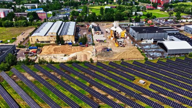 Aerial view of a solar farm with rows of photovoltaic panels adjacent to an industrial facility with warehouses and construction materials in Latvia.