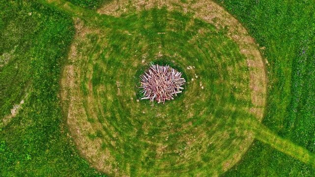 Aerial view of a circular pattern mowed into a vibrant green meadow with a central pile of stacked wood in Latvia.