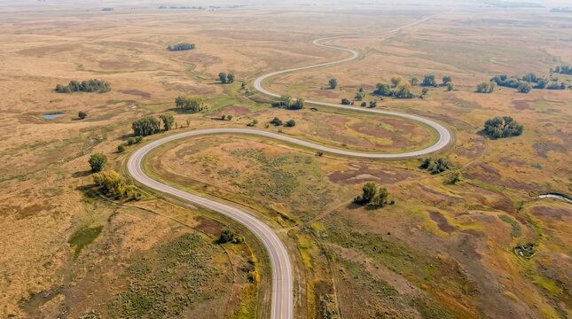 Serpentine Road Through Vast Plains: An aerial view captures a winding road snaking through dry, golden plains dotted with sparse vegetation and small groves of trees, under a hazy sky.