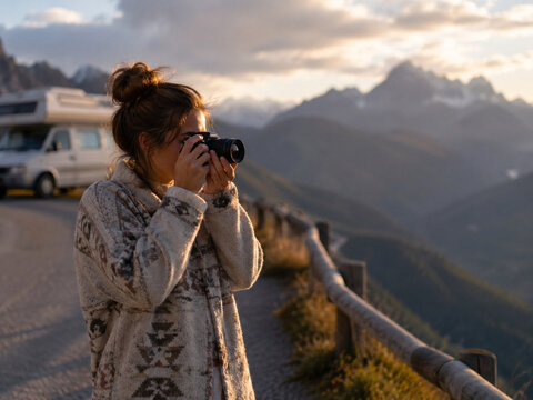 Female traveler photographing dramatic mountain peaks at golden hour beside a parked camper, with the alpine viewpoint softly blurred behind.