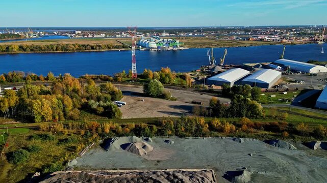 Aerial view of the industrial area of Riga Port with storage warehouses, piles of gravel, and the Daugava River under a clear sky in Riga, Latvia.
