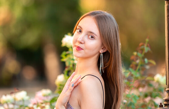 Elegant young woman posing among blooming pink roses in a summer garden. Romantic beauty portrait with long hair, stylish outfit and natural floral background, lifestyle concept.