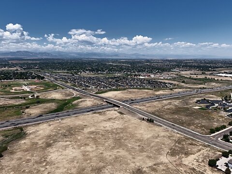 Aerial view of a highway intersection surrounded by suburban residential neighborhoods and open fields under a clear blue sky in Thornton, Colorado, United States.